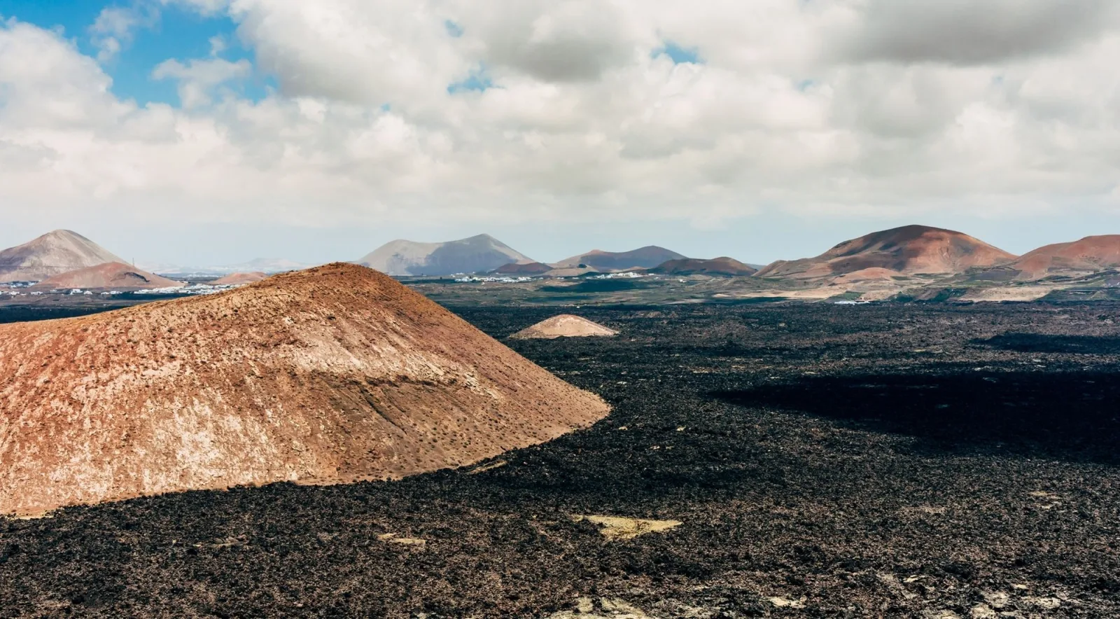A rust-coloured volcanic cone rising from a field of black lava, with more cones on the horizon