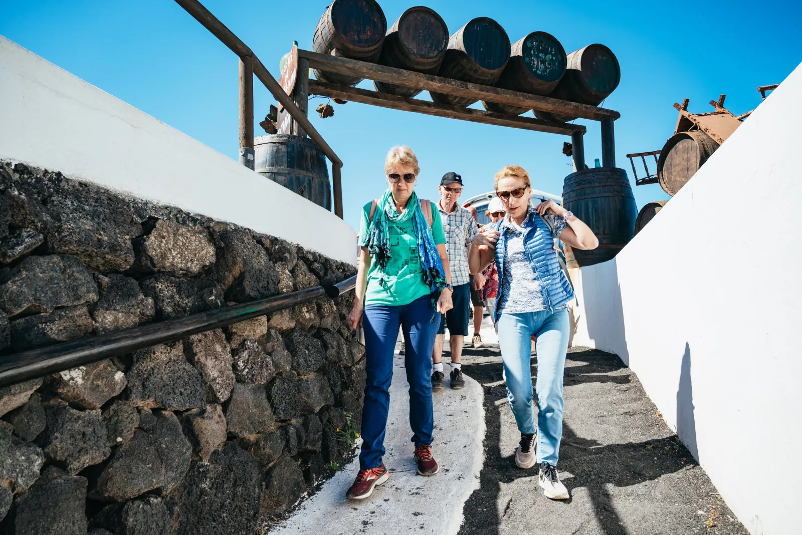 Two visitors walking a whitewashed ramp past a lava-stone wall hung with barrels