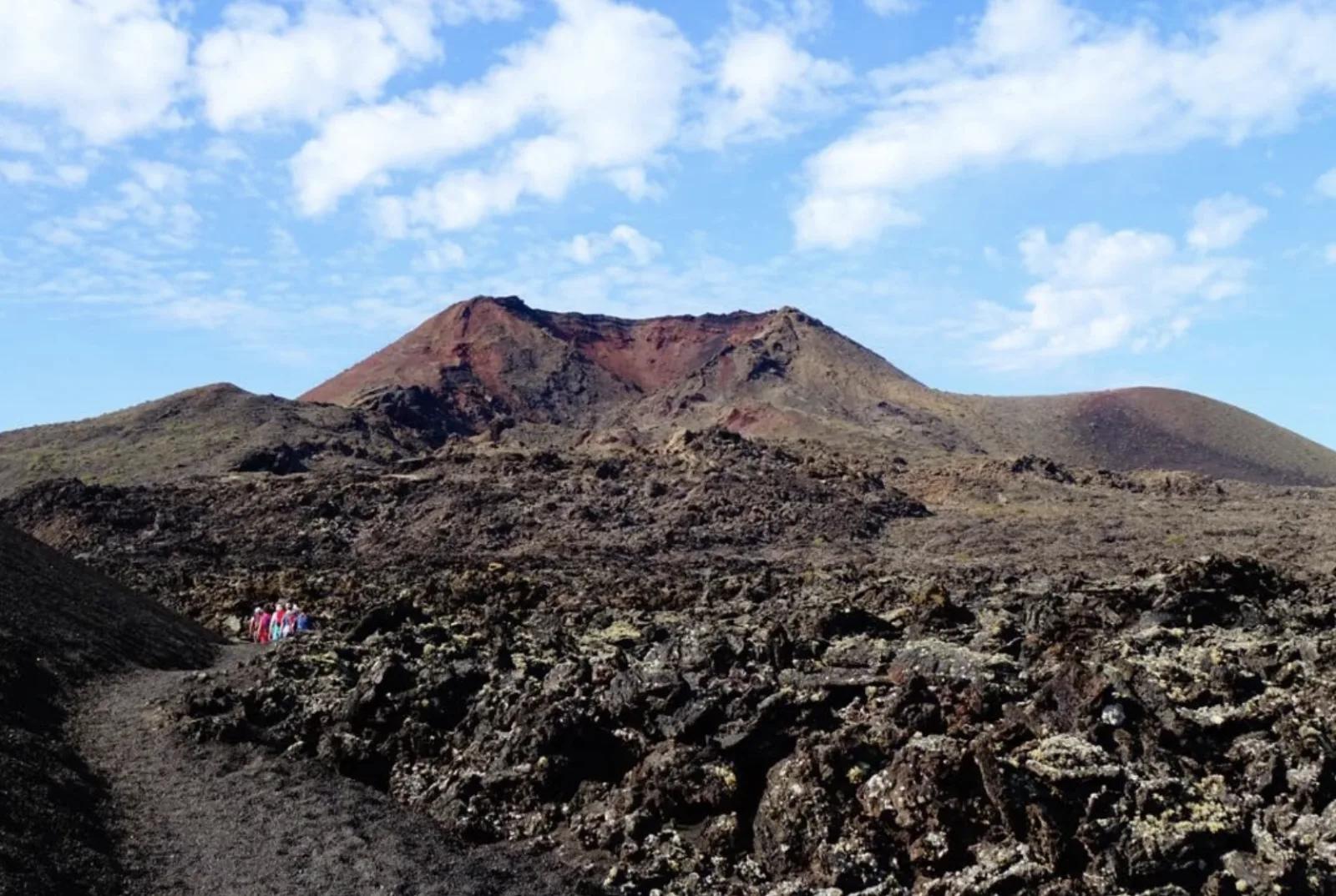 A red-rimmed crater rising above a broken black lava flow under a cloud-streaked sky