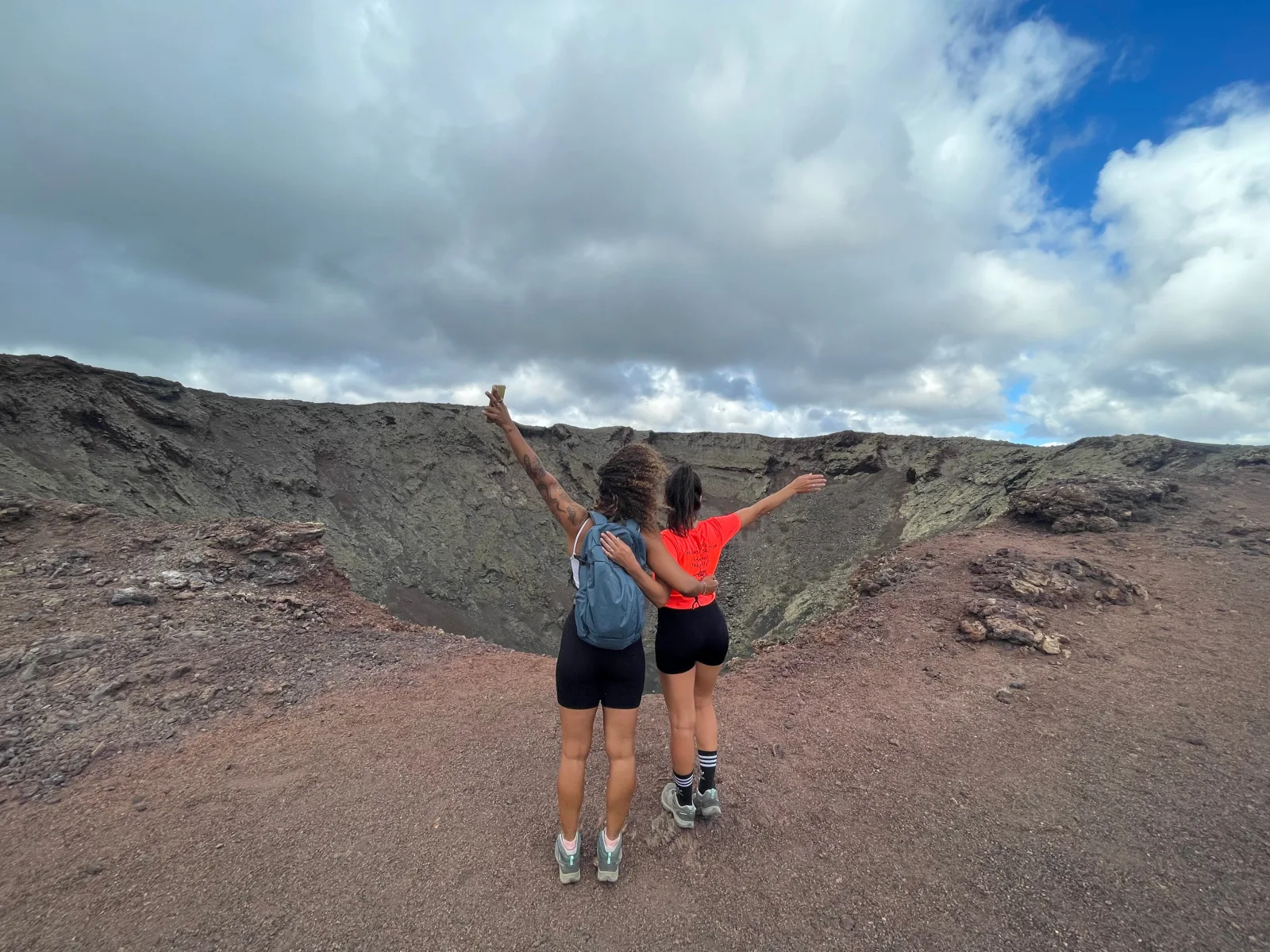 Two hikers standing arms-wide on the rim of a wide crater under a cloudy sky