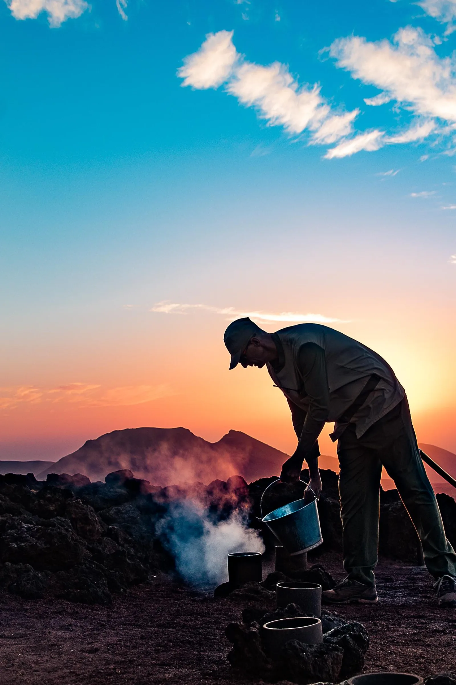 A cook grilling over the open geothermal heat vent as the sun drops behind the cones